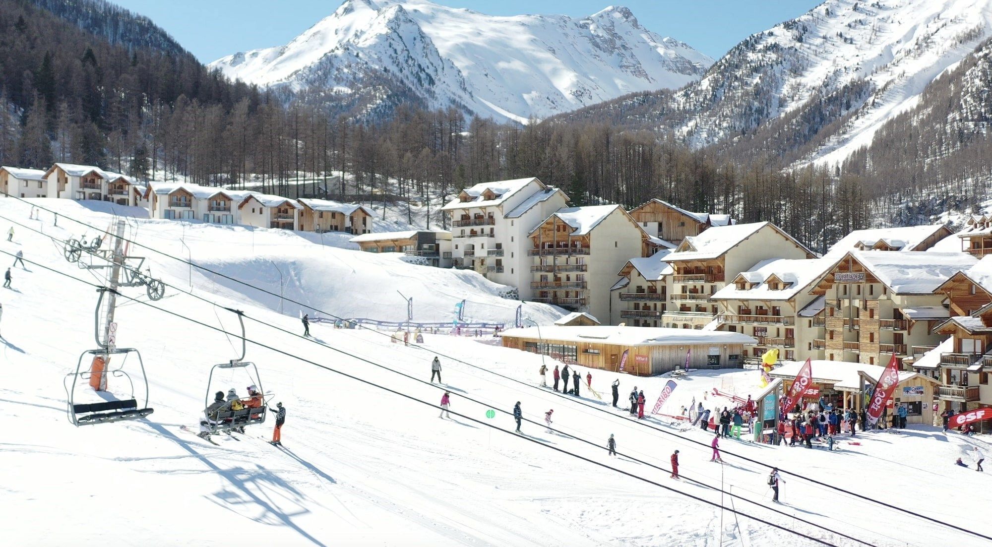 A ski resort with multiple chalet-style buildings, a ski lift, and a snowy mountain backdrop.