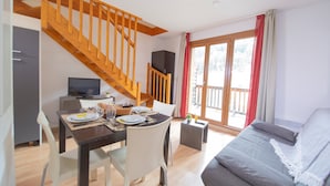 Un salon moderne avec un canapé gris, un escalier en bois, un coin repas avec une table dressée pour deux personnes et une grande fenêtre ornée de rideaux rouges.