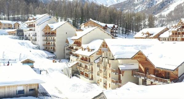 A snow-covered resort with multiple wooden buildings, balconies, and a ski slope in the background.