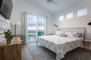 A bedroom with a bed, a desk, a chair, a TV, and a view of the sea.