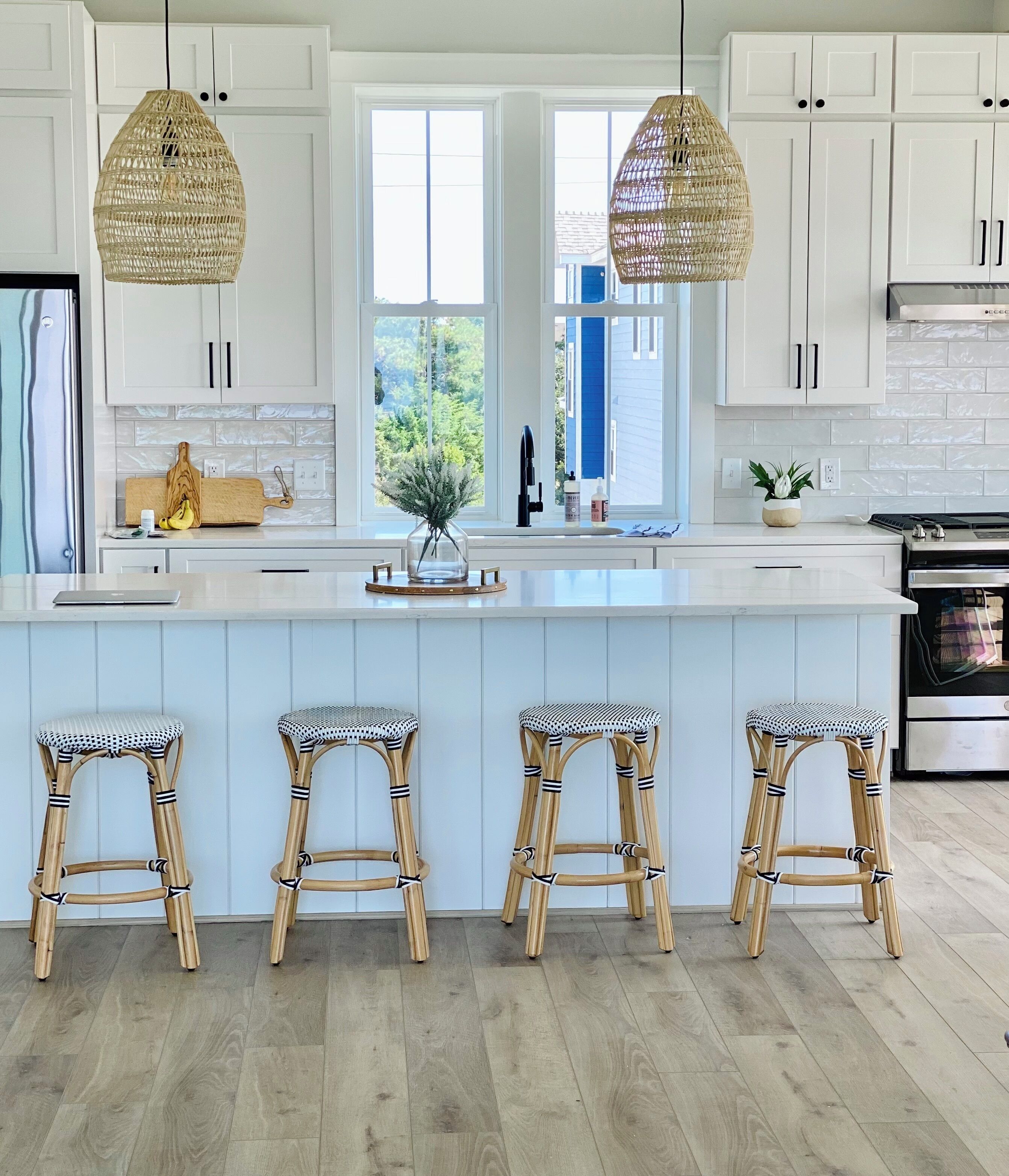 A modern kitchen with white cabinets, a large island with stools, and hanging rattan light fixtures.