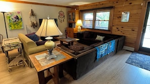 A cozy living room with a wooden floor, a sofa, a table with a lamp, and a television. - Luxury Log Cabin with work spaces, generator and car charger  (Tiny Township)