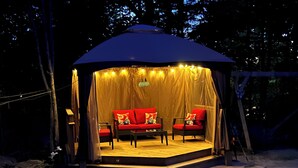 A tented outdoor seating area with red sofas and a wooden table, surrounded by trees. - Luxury Log Cabin with work spaces, generator and car charger  (Tiny Township)