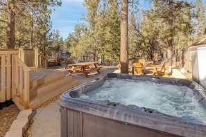 A hot tub surrounded by wooden deck steps, picnic tables, and tall pine trees.