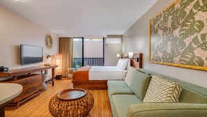Habitación de hotel con cama, sofá verde, mesa de centro de madera, televisor y un cuadro grande con motivos florales en la pared.