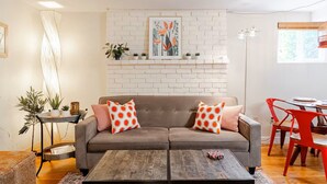 A living room with a brown sofa, two orange polka dot pillows, a wooden coffee table, a red chair, and a white brick wall with a framed botanical print. - Spacious 1 bedroom Ballard Apartment, Walk to Dining & Shops,  15 Min to Seattle (Seattle)