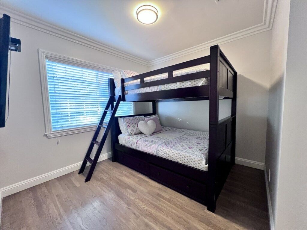 Een stapelbed met een ladder, een raam met zonwering en een lamp aan het plafond.