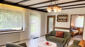 Una sala moderna con sofá, sillones y una mesa de centro. Se aprecia una ventana con cortinas translúcidas, una puerta de madera y un estante con objetos decorativos.