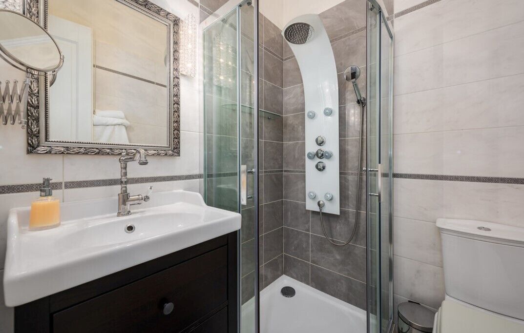 Un baño moderno con ducha de vidrio, un lavamanos blanco con dispensador de jabón y un espejo grande con marco decorativo.