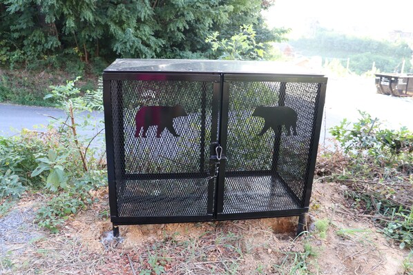 Una caseta de metal negro con siluetas de dos animales, rodeada de follaje natural y una carretera al fondo.