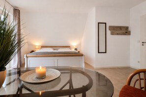 A bedroom with a bed, bedside lamps, a bedside table with a candle, a potted plant, and a wall-mounted picture frame.