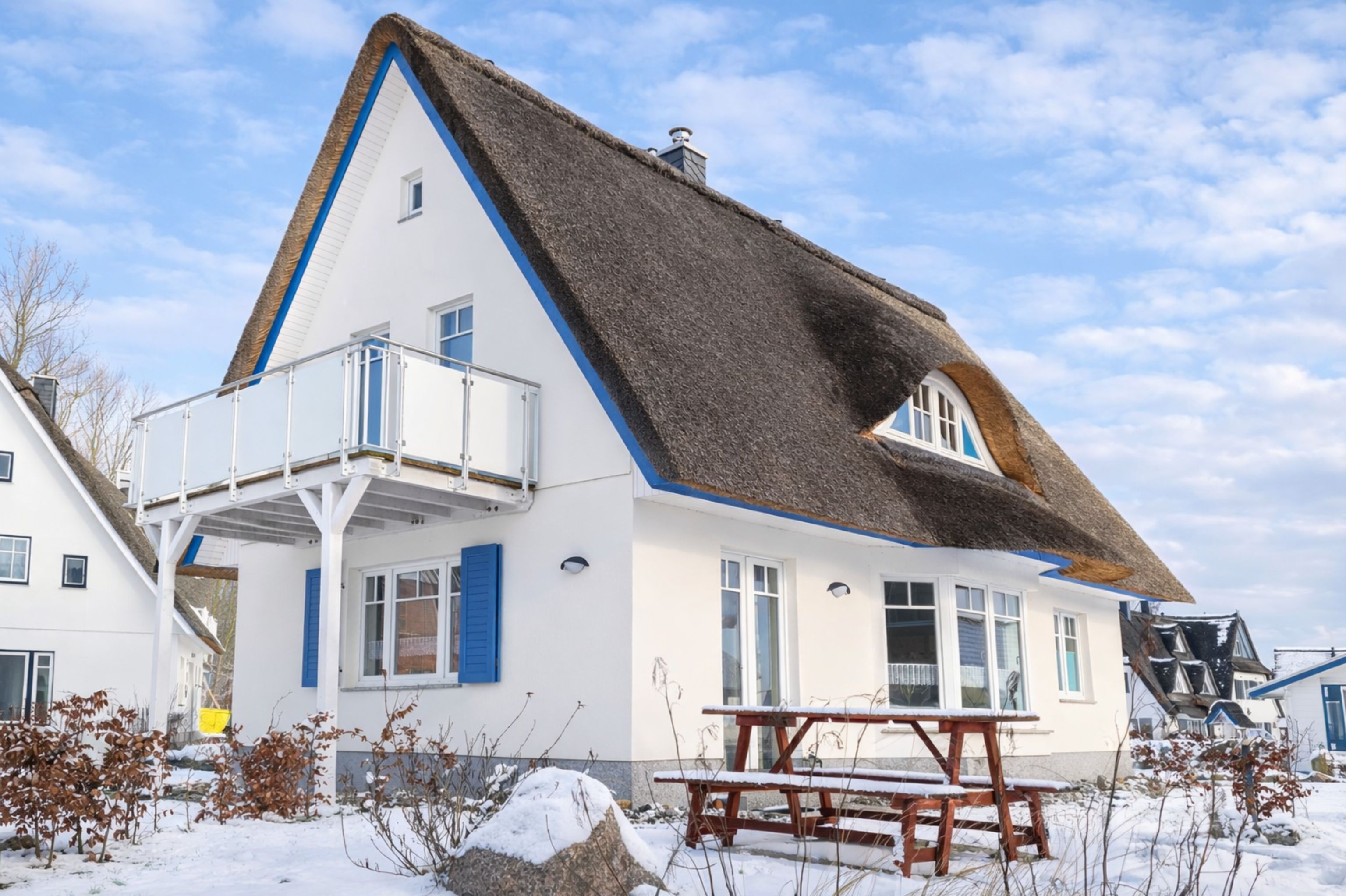 Une maison blanche avec un toit de chaume, un balcon et un banc en bois au premier plan.