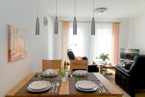A dining area with a wooden table set for two, a sofa, a TV, and a painting on the wall. - 1.1 - Glienbergweg 26 Zinnowitz (Zinnowitz)
