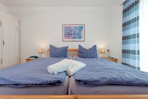 A double bed with a blue bedspread and pillows, a bedside table with a lamp, a framed abstract painting on the wall, and a window with blue curtains.