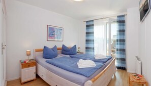 A double bed with blue bedding, a bedside table with a lamp, a framed picture on the wall, and a window with curtains. - 1.1 - Glienbergweg 26 Zinnowitz (Zinnowitz)