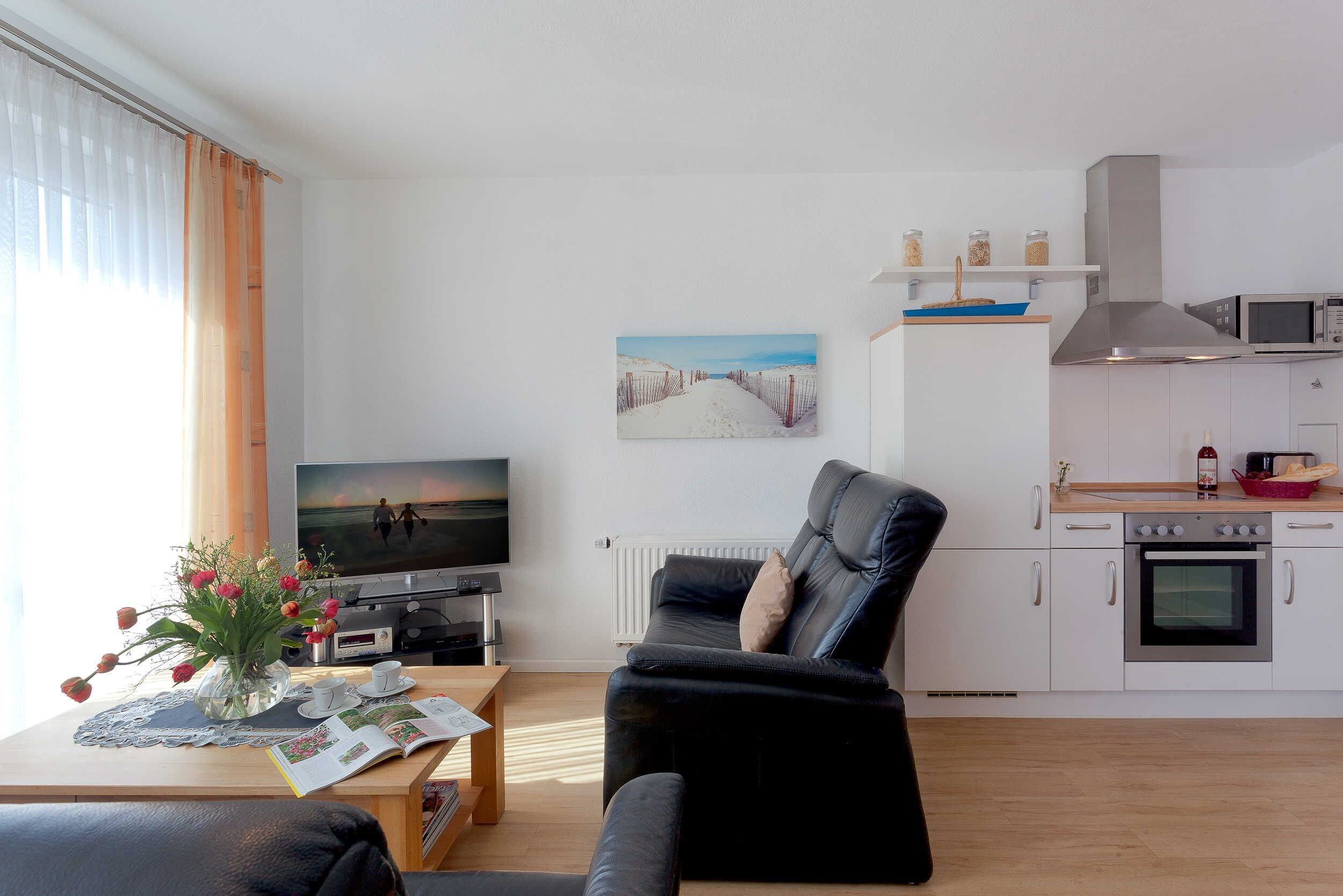 Un soggiorno moderno con una TV a schermo piatto, un divano in pelle nera, un tavolo da pranzo con un vaso di fiori e un angolo cottura con pensili bianchi e un forno a microonde.