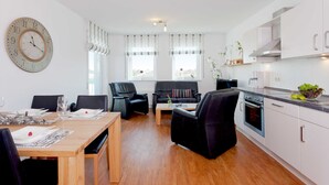 Ein modernes Wohnzimmer mit Esstisch, schwarzen Ledersofas und einer Küchenzeile mit weißen Schränken und einem eingebauten Backofen.