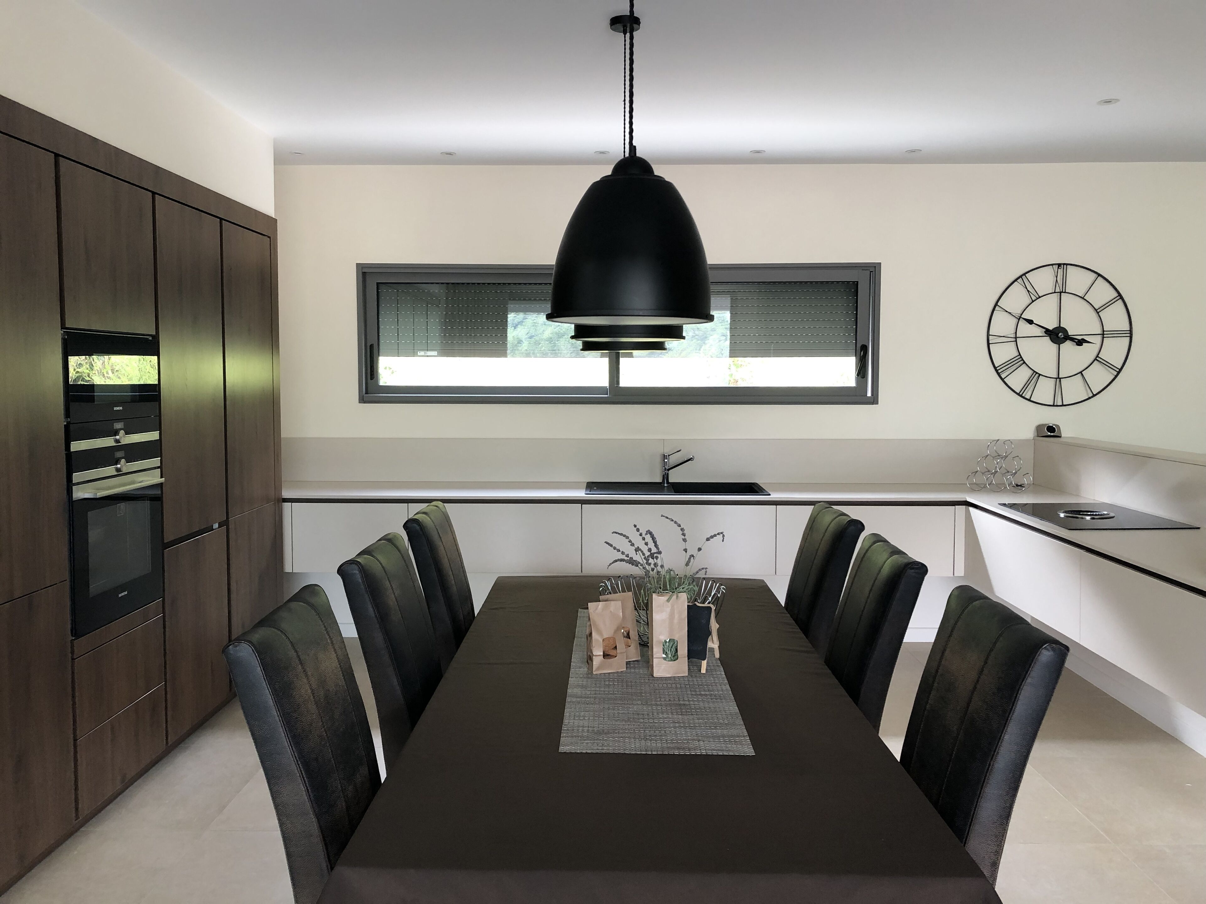 Una cocina moderna con comedor, que incluye una mesa grande, sillas, una lámpara colgante, una ventana y un reloj de pared.