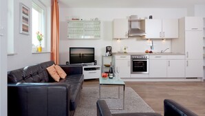 A modern living room with a black leather sofa, a glass coffee table, and a flat-screen TV. Adjacent is a kitchen with white cabinets, a built-in oven, and a sink. - 2.3 - Glienbergweg 26 a Zinnowitz (Zinnowitz)