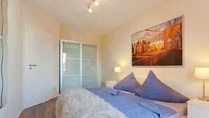 A bedroom with a bed, a nightstand, a lamp, a painting, and a door. - 2.3 - Glienbergweg 26 a Zinnowitz (Zinnowitz)