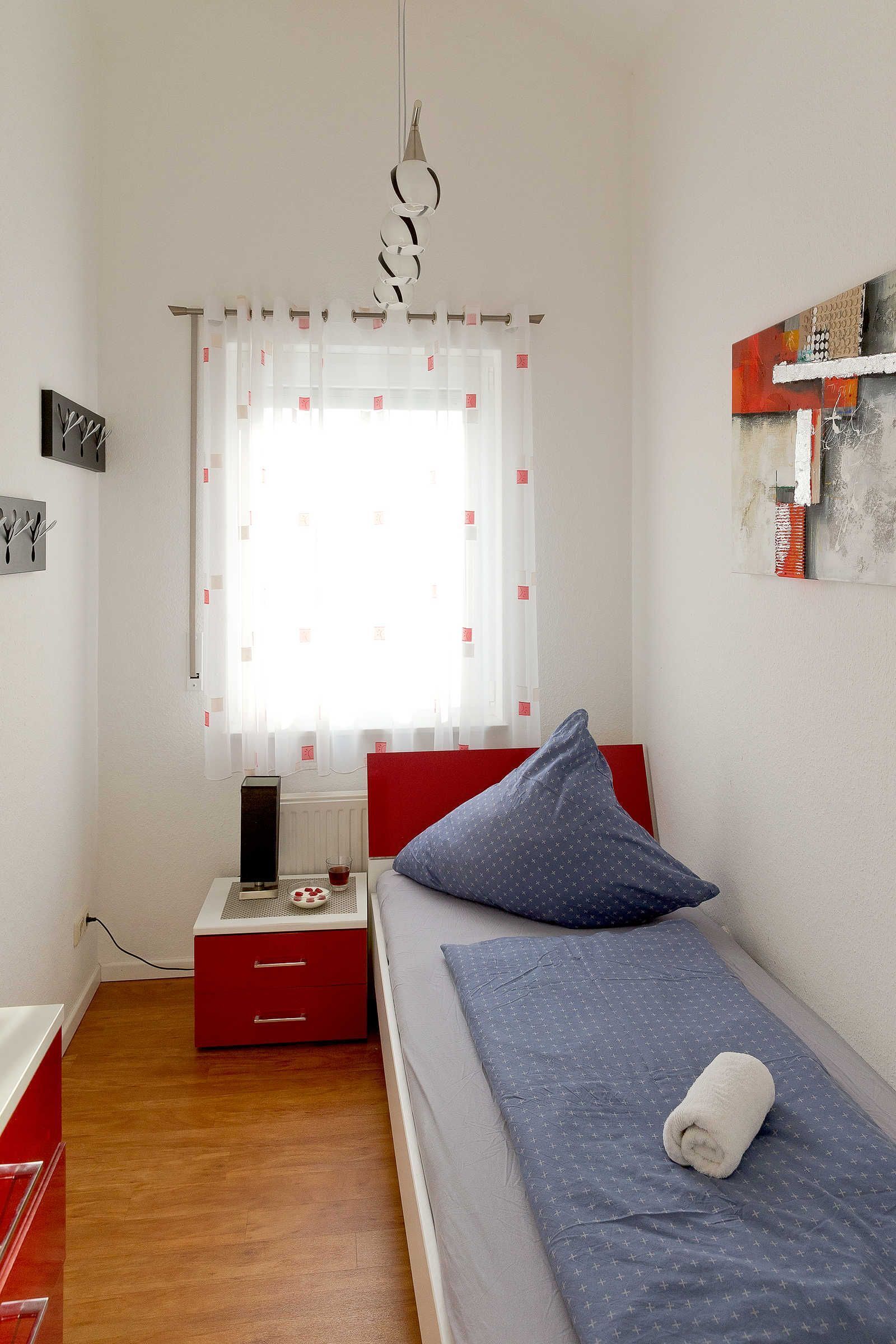 A small, neatly arranged bedroom with a single bed, a red nightstand, a white radiator, and a window with sheer curtains.