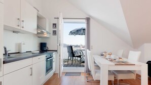Ett modernt kök med vita skåp, en matplats med ett vitt bord och stolar, och utsikt över utomhusmiljön genom en skjutdörr.