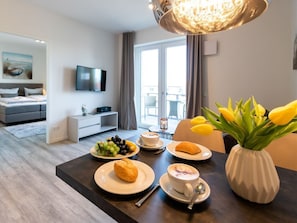 A modern hotel room with a dining area, a flat-screen TV, a large window, and a dining table set with breakfast.