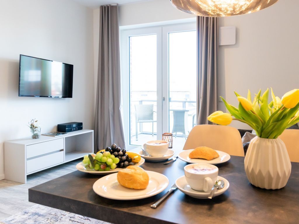 A hotel room with a dining table set with breakfast, a flat-screen TV mounted on the wall, and a view of the outdoors through the window.