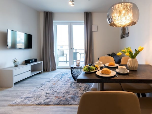 Une chambre d’hôtel moderne dotée d’une télévision à écran plat, d’une table à manger avec des produits pour le petit-déjeuner, d’un grand lustre et d’une vue sur l’extérieur, visible à travers les portes coulissantes en verre.