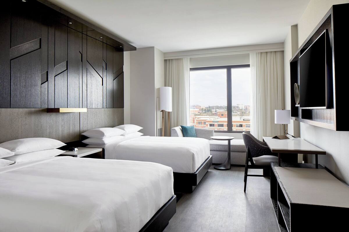 A hotel room with two beds, a desk, a chair, a TV, and a large window with a city view.