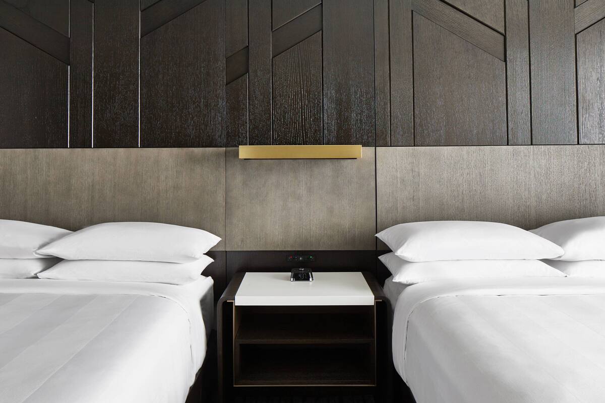 A hotel room with two beds, a bedside table, a nightstand, and a wall-mounted light fixture.