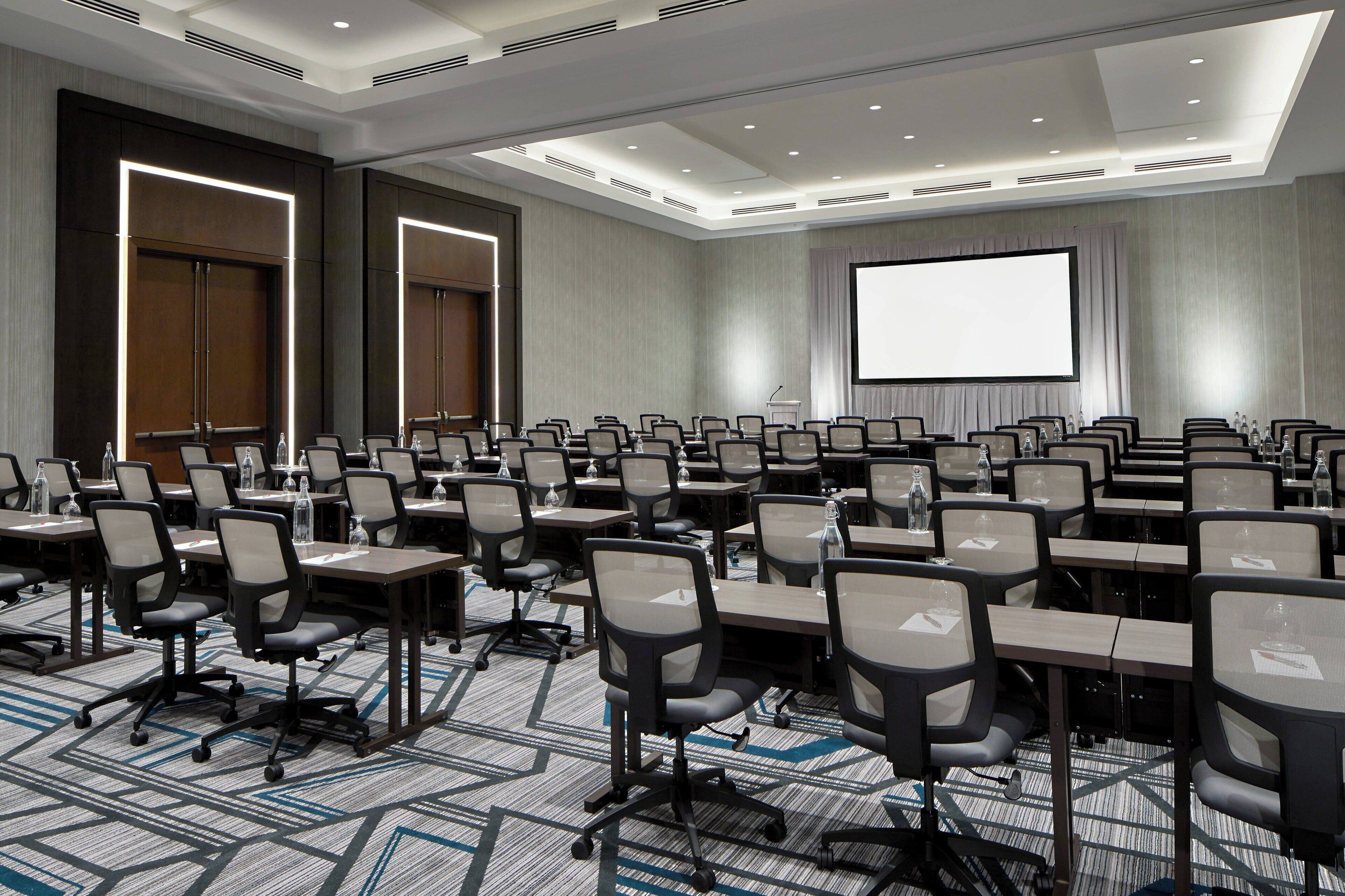A conference room with rows of tables and chairs, a large screen, and a podium.