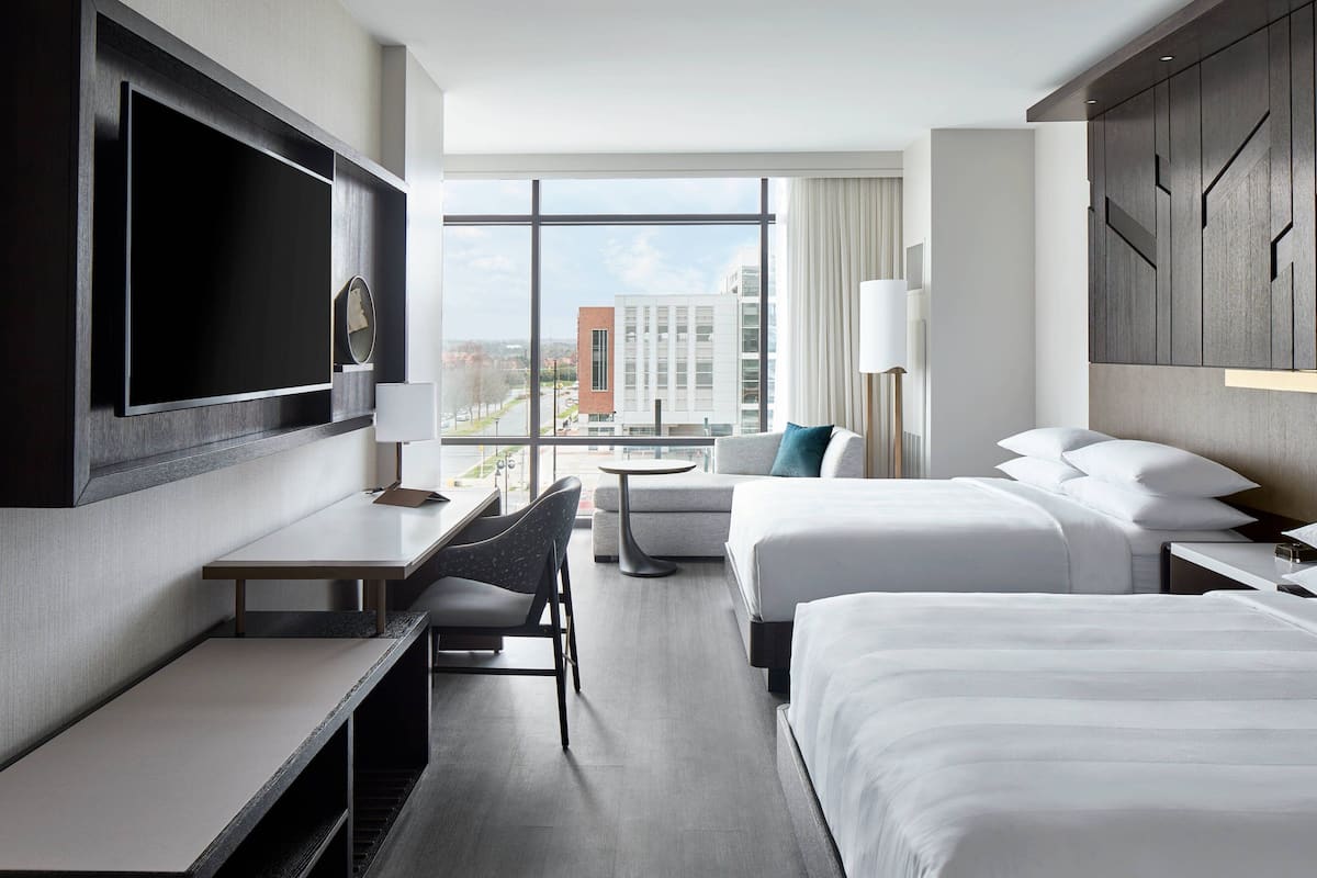 A hotel room with a large TV, a desk, two beds, a chair, and a view of the city.