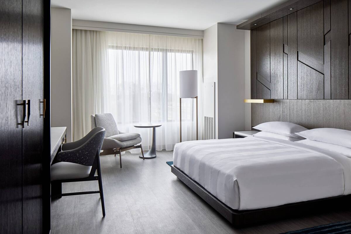 A modern hotel room with a large bed, a desk, a chair, and a floor lamp.