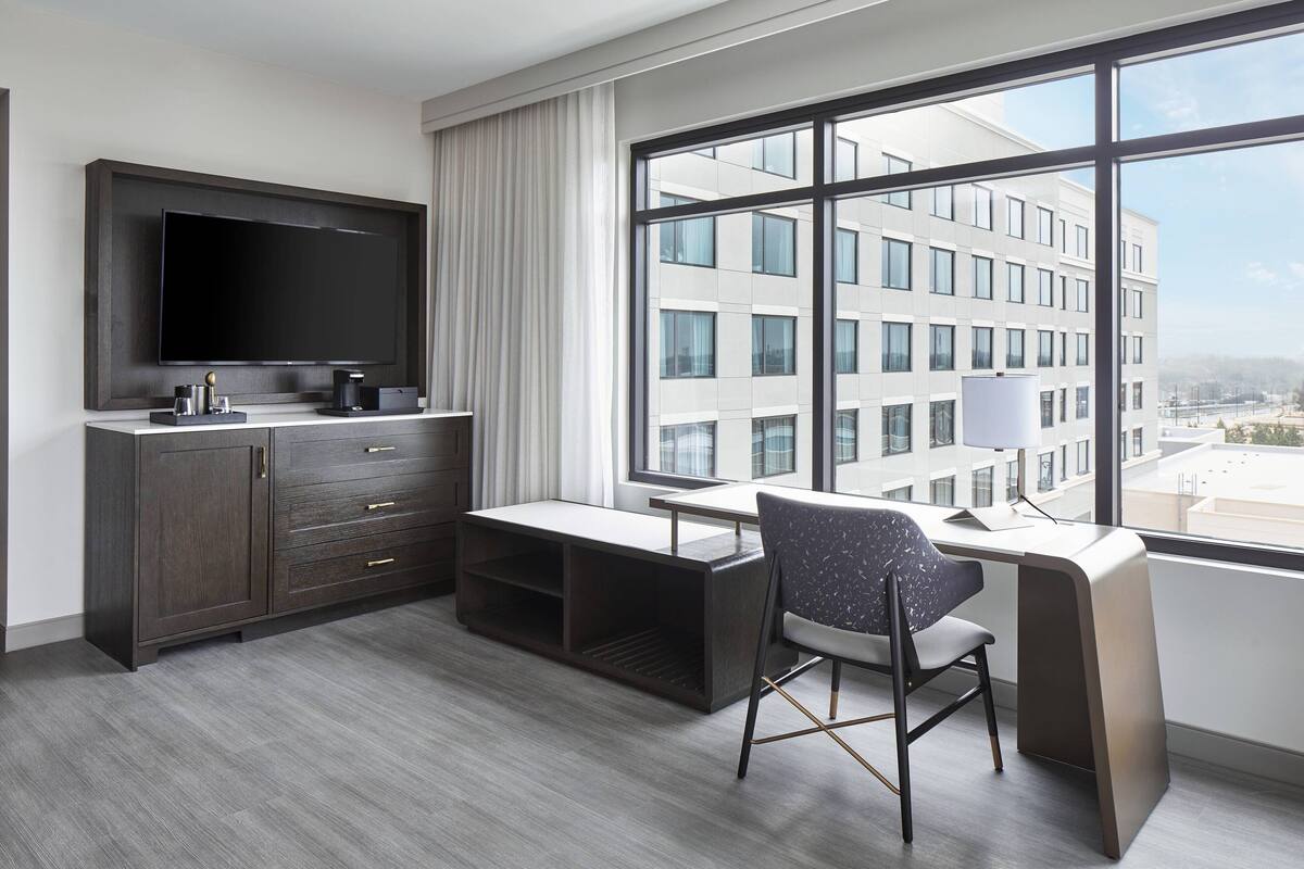 A modern hotel room with a large window, a flat-screen TV, a desk, and a chair.