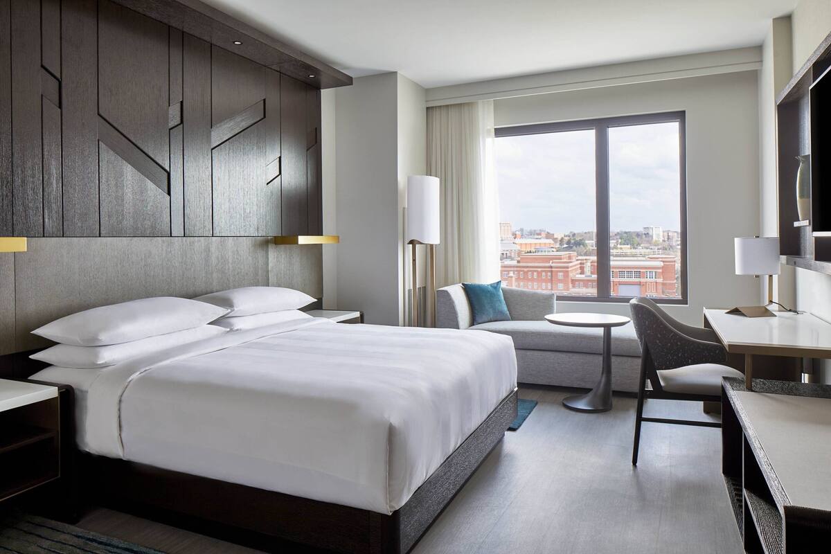 A modern hotel room with a large bed, a sofa, a desk, and a view of the city.