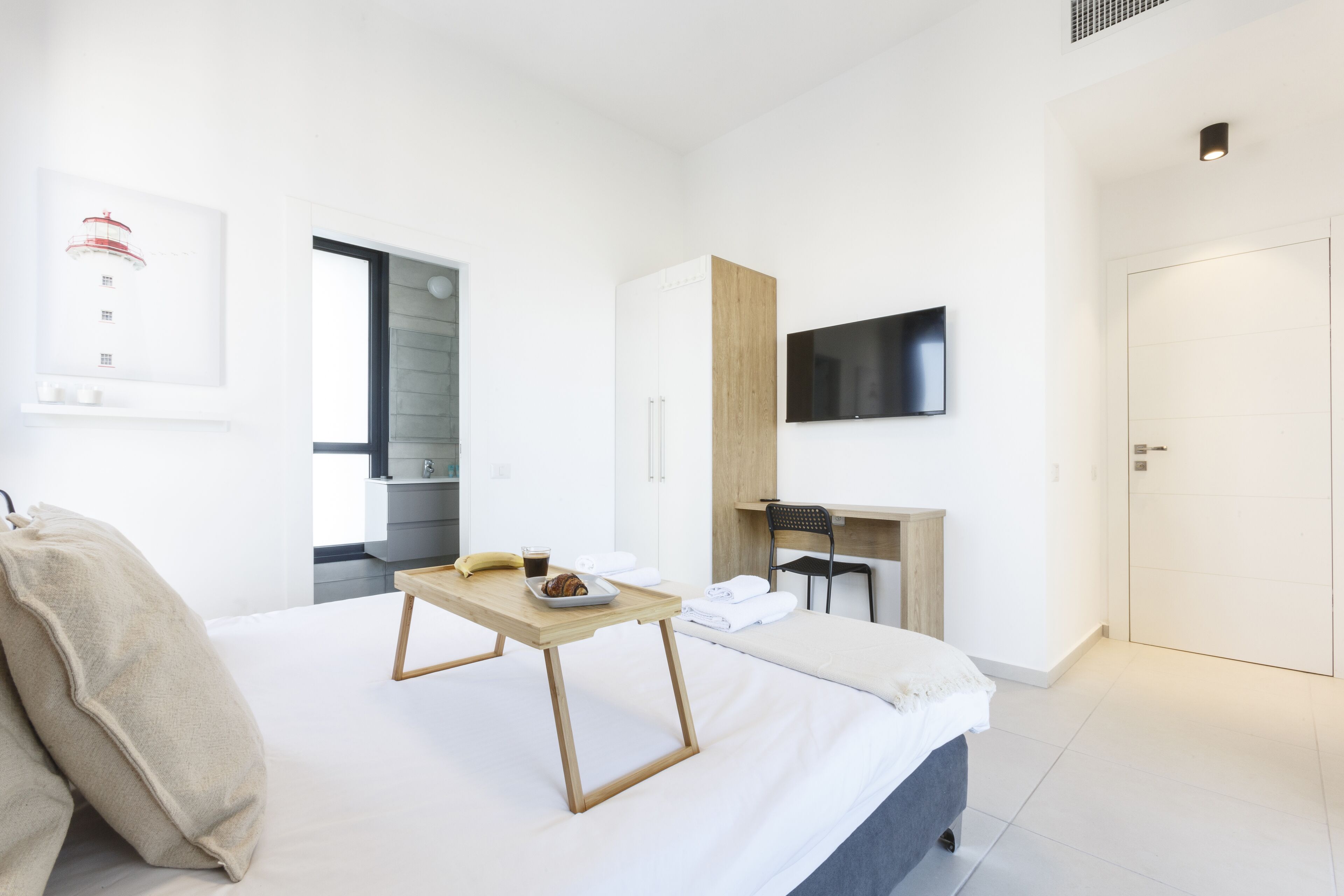 A modern hotel room with a bed, a wooden bedside table with a tray of breakfast, a TV mounted on the wall, and a door leading to the bathroom.