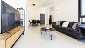 Una sala de estar moderna con televisor de pantalla plana, un sofá de color oscuro, una mesa de centro y un comedor con sillas y una mesa.