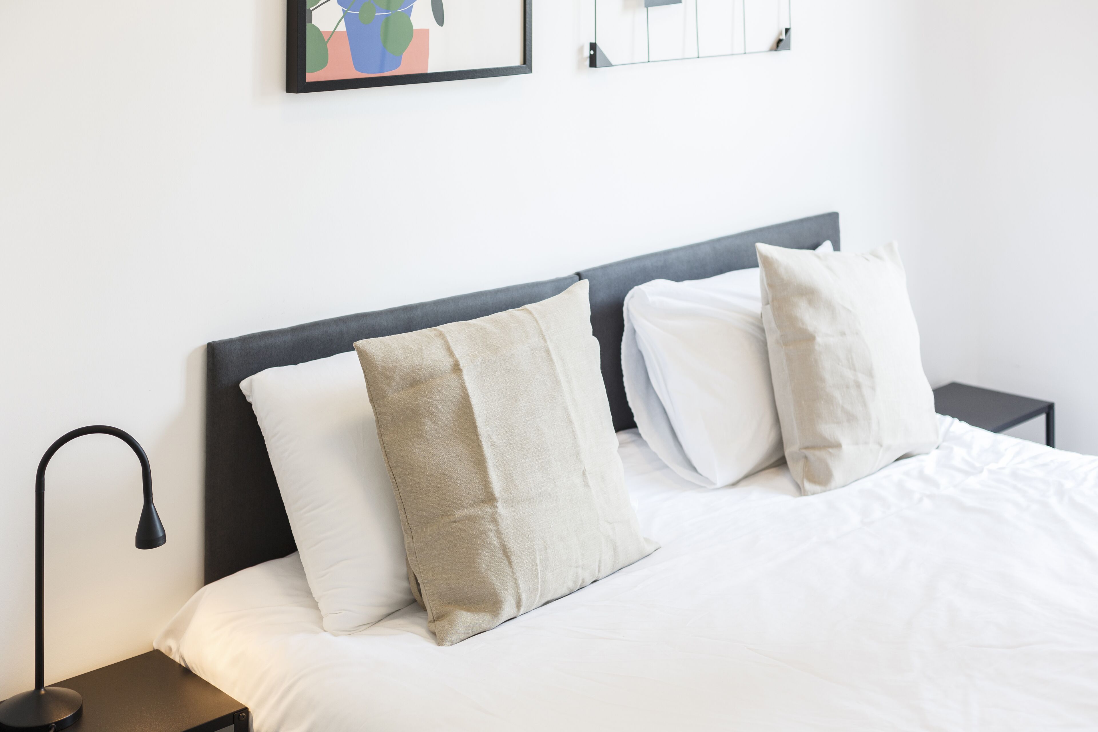 A neatly made bed with white bedding and two pillows, a bedside table with a lamp, and a wall-mounted artwork above the bed.