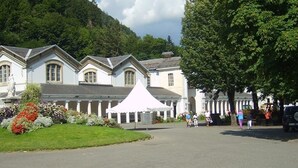 A white building with a central pavilion, surrounded by a well-maintained garden and a mountain backdrop.