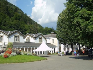Un edificio bianco con un corpo centrale, circondato da un giardino curato e con le montagne sullo sfondo.