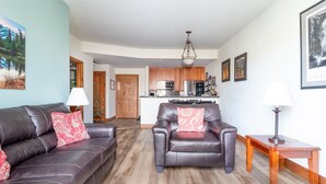 A living room with a brown leather sofa and armchair, a wooden coffee table, and a kitchen in the background.