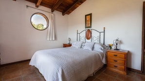 Quarto com cama, cabeceira de madeira, mesas de cabeceira, um espelho redondo e um quadro na parede.