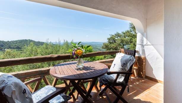 Uma varanda com mesa e cadeiras de madeira, um vaso com flores e vista para colinas verdes.