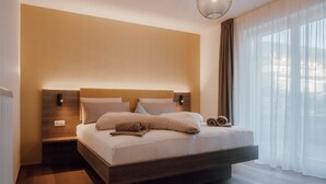 A modern bedroom with a large bed, wooden headboard, and a city view through sheer curtains. - Apartment Kronsun "Joy" – Directly at the Kronplatz Valley Station (Percha)