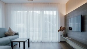 A modern living room with a sofa, a coffee table, a flat-screen TV mounted on the wall, and sheer curtains. - Apartment Kronsun "Joy" – Directly at the Kronplatz Valley Station (Percha)