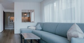 A modern living room with a grey sofa, a small wooden table, and a kitchen area with a sink and cabinets.