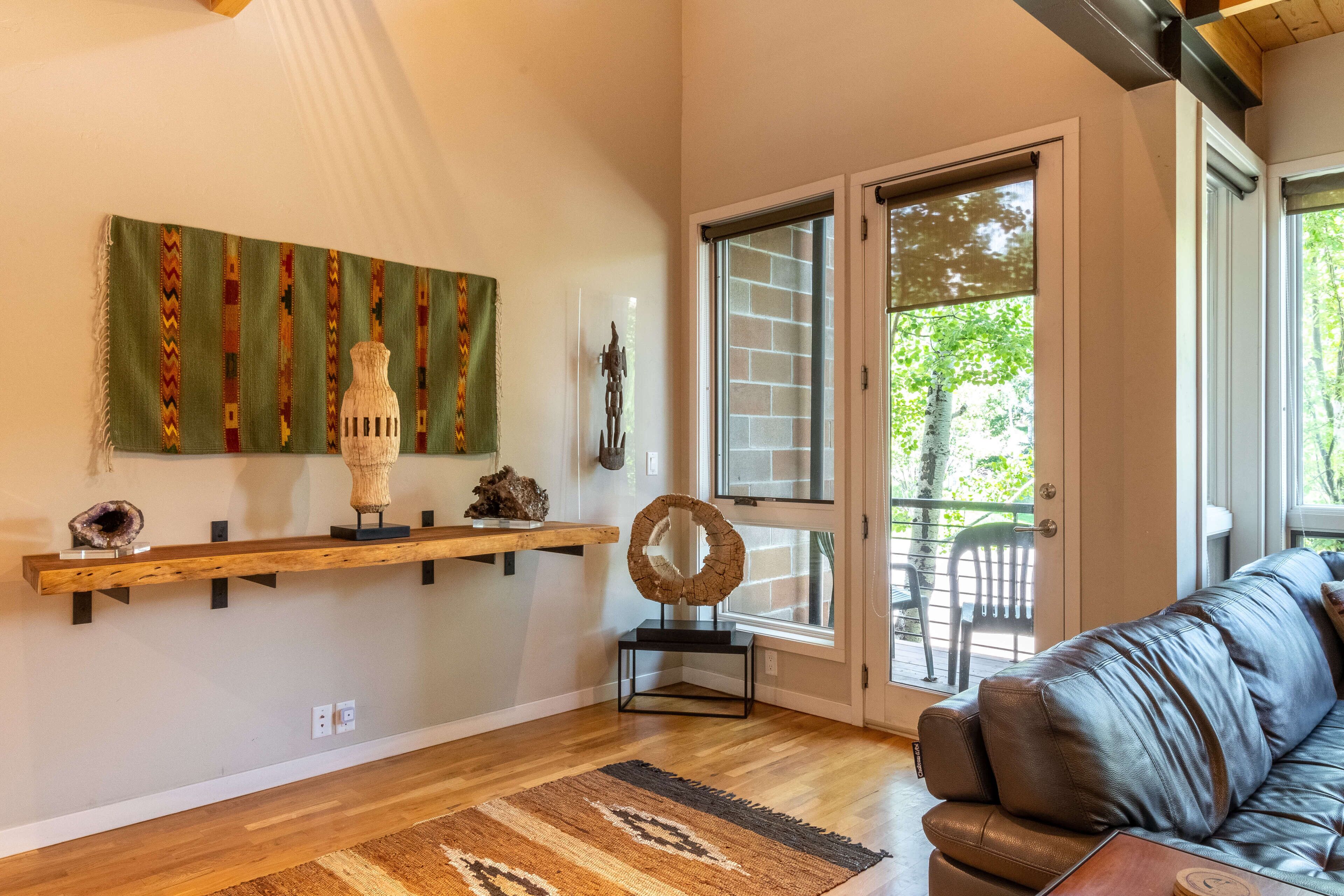 A living room with a wooden floor, a brown leather sofa, a green wall hanging, and a glass door leading to an outdoor area.