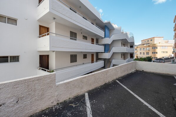 A multi-story residential building with balconies, a parking area, and a clear sky.