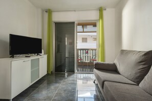 A living room with a sofa, a television on a cabinet, a sliding glass door, and a balcony with a table and chairs.